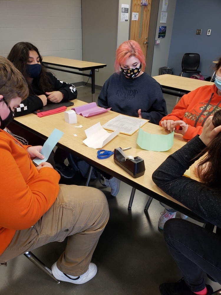 students sit around a school table - with masks on- working on their Econ project using paper, pencil, and scissors 