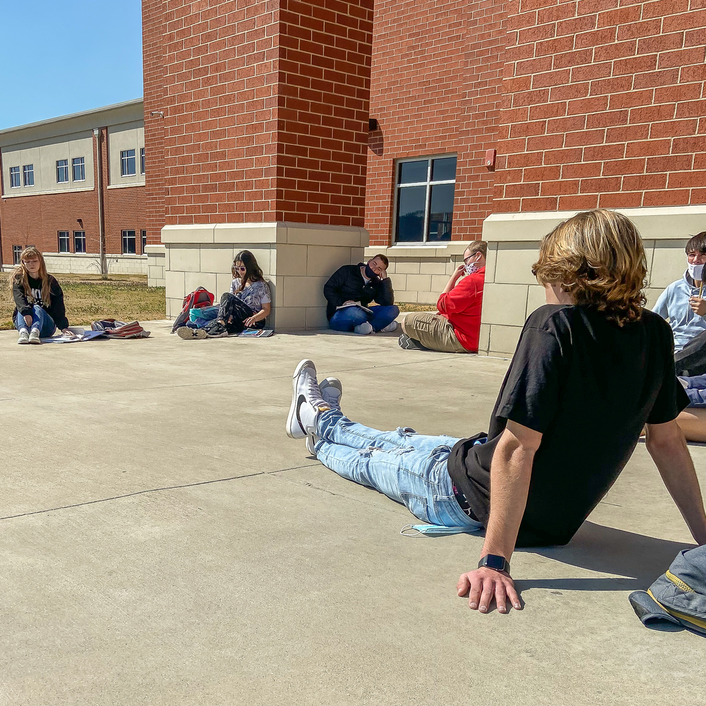 6 students sit outside the high school front doors in a social distanced circle discussing